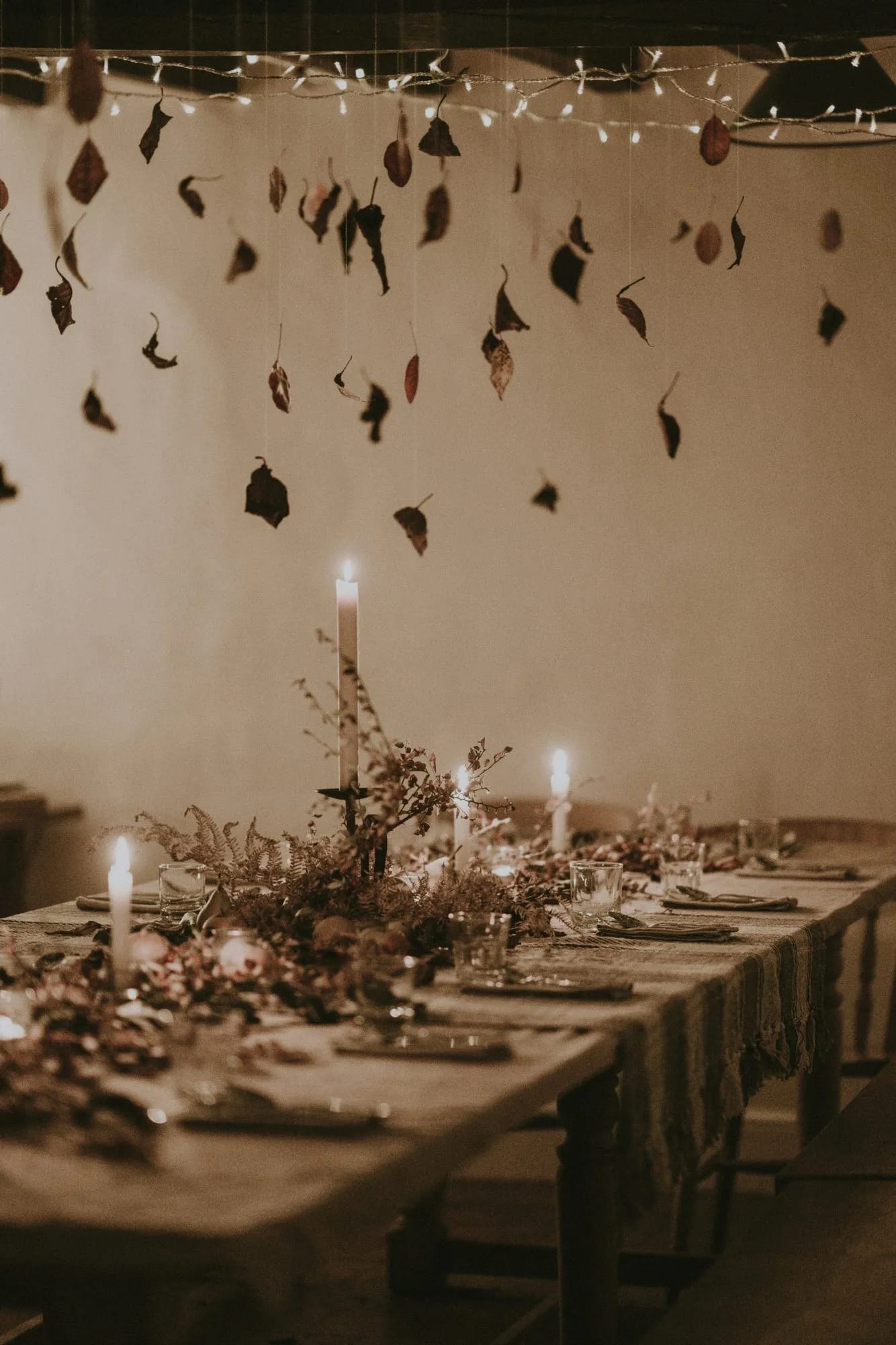 A long linen-covered table with candles and autumn foliage beneath hanging leaves