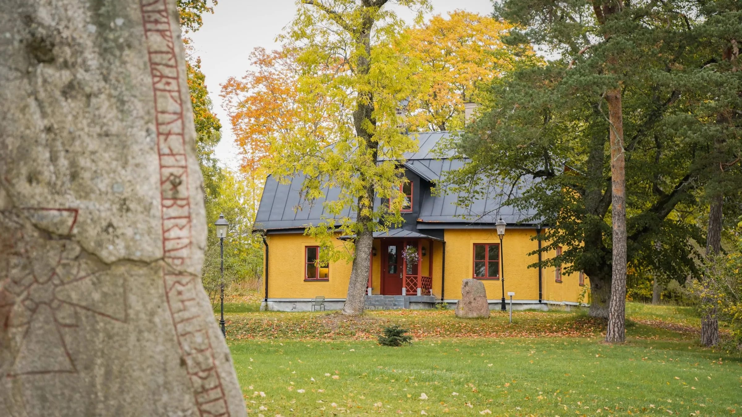 A runestone in the foreground with the wing at Hagby behind autumn trees